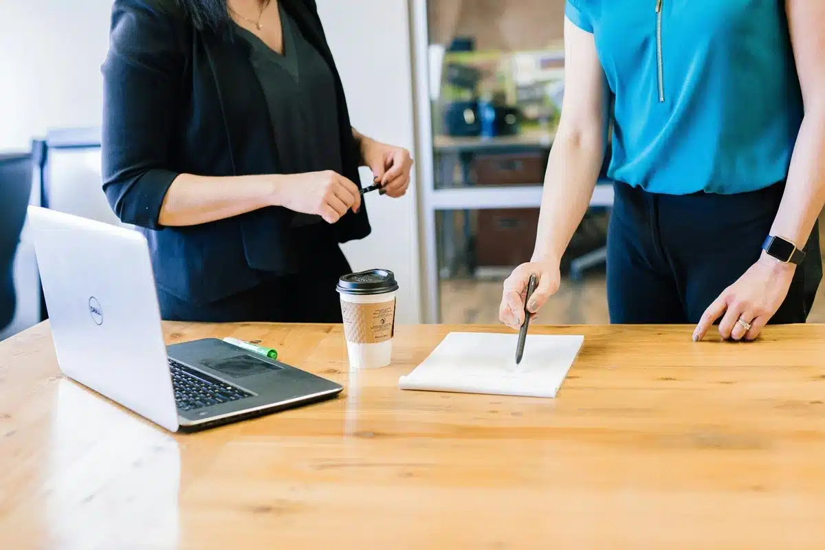 Vue du buste de 2 femmes en train de parler d'un sujet en prenant des notes sur un cahier posé sur une table avec un ordinateur.